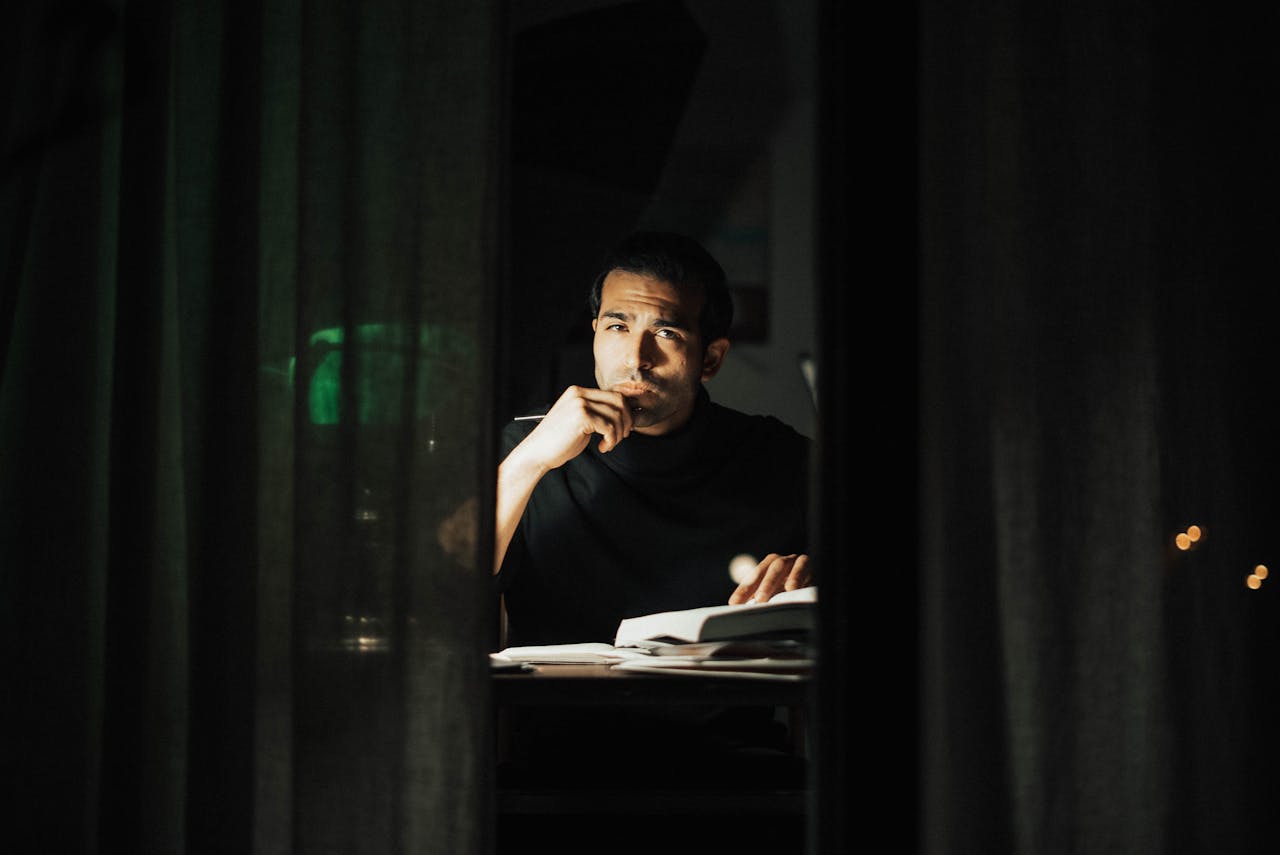 Serious young male wearing black turtleneck reading book and looking at camera while touching chin in contemplation and sitting at desk in dark home office
